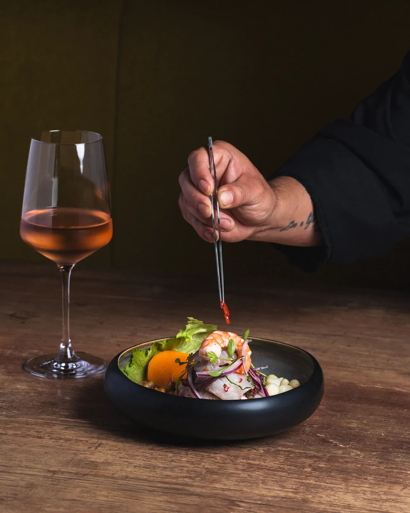 A chef plating a Pachamama ceviche with tweezers, a glass of wine beside it