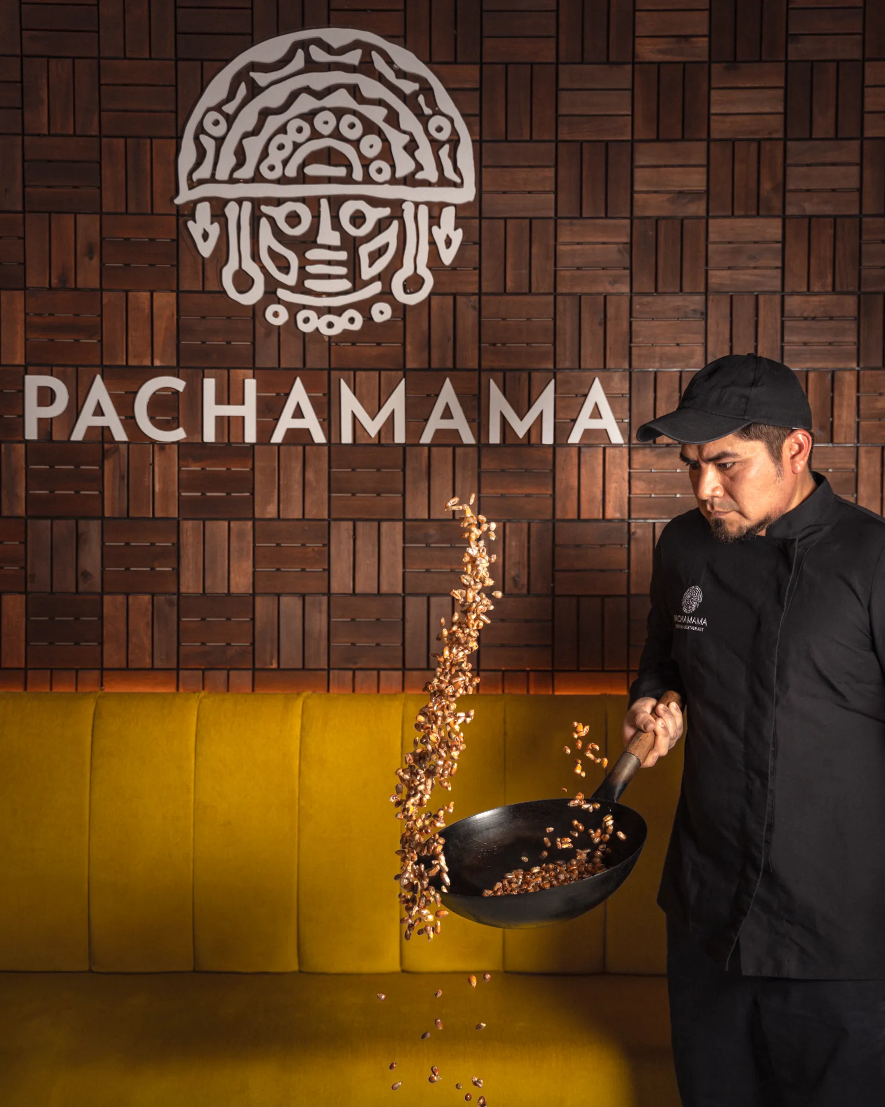 A Pachamama chef tossing toasted cancha in a pan beneath the carved mask on the dining room wall