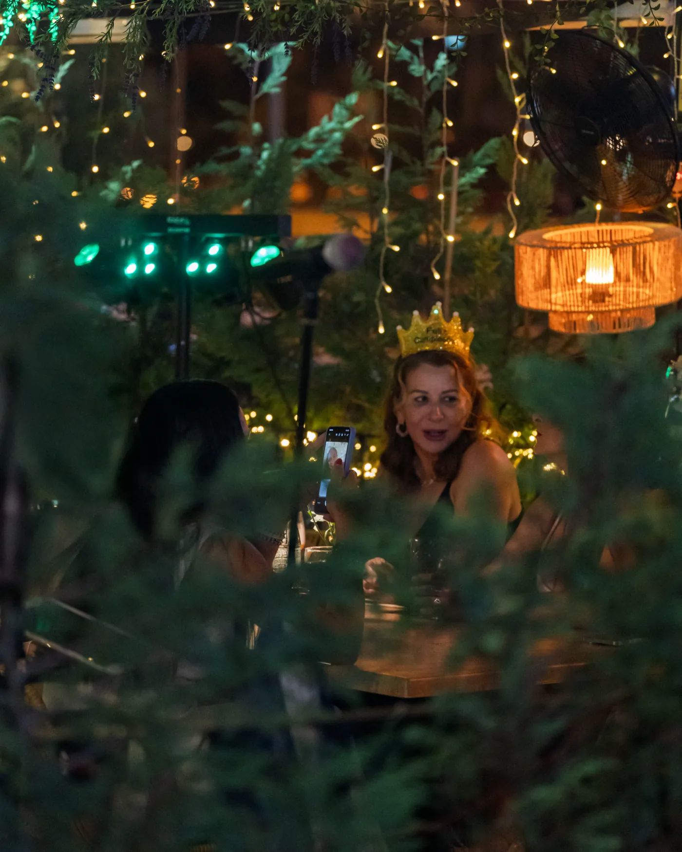 A guest in a gold birthday crown being filmed by friends under the string lights at Pachamama